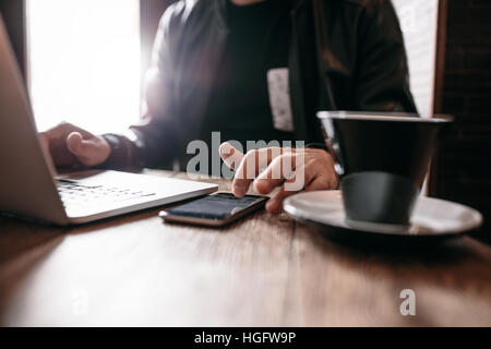 Immagine ravvicinata di uomo seduto presso il cafe con il computer portatile utilizzando il telefono cellulare. Le mani dell'uomo utilizzando smart phone al coffee shop Foto Stock