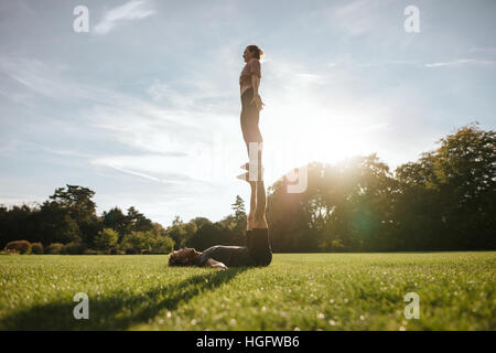 Montare il giovane facendo acrobatici esercizi di yoga nel parco. Uomo disteso su erba e donna di bilanciamento sui suoi piedi. Foto Stock