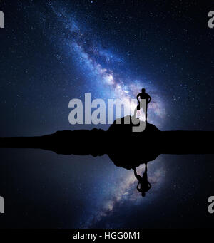Via Lattea. Di notte il cielo stellato e la silhouette di un uomo in piedi con zaino sulla pietra vicino al lago Foto Stock