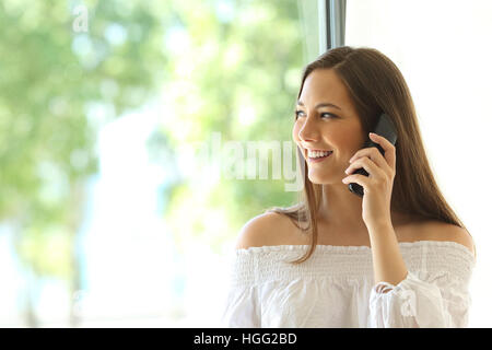 Ragazza chiamata sul telefono fisso e guardando all'esterno attraverso una finestra nel salotto di casa Foto Stock