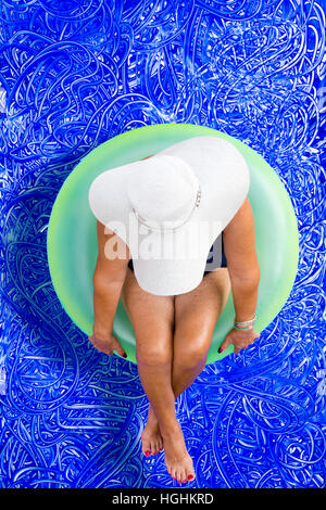 Conciate donna godendo una giornata in piscina per la sua vacanza estiva seduta galleggiante su un verde del tubo di plastica che indossa un ampio ed elegante cappello visto da abo Foto Stock