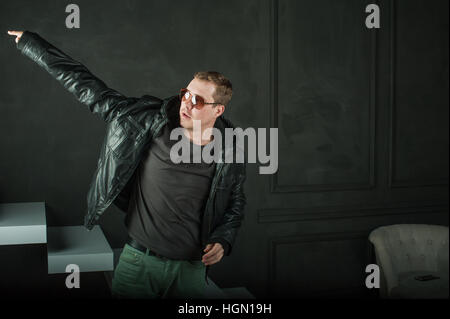 Studio fotografico giovane ragazzo brutale. l uomo in occhiali da sole, T-shirt, jeans mette nero su giacca di pelle. Sfondo nero pareti dipinte di bianco e scale Foto Stock