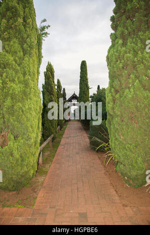 Alte siepi di giardino che conduce al mandrino in legno al tramonto con il marciapiede di mattoni in diminuzione in prospettiva. Foto Stock