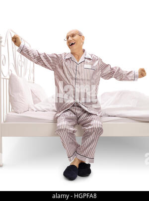 Uomo anziano in pigiama seduto su un letto e sbadigliamento isolati su sfondo bianco Foto Stock