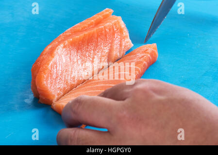 Close up di cuochi mani affettatura di filetto di salmone fresco longitudinalmente sulla superficie blu Foto Stock