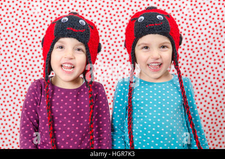 Due gemelle sono sorridente alla fotocamera e di essere felice. I bambini piccoli sono gioiosa. Bambino sta indossando un cappello di lana a forma di coccinella su un backgrou Foto Stock