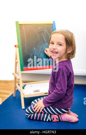 Piccolo bambino è il disegno con pezzi di Colore gessetto su chalk board. Ragazza è esprimere la creatività e guardando la telecamera, sorridente in un nido, Foto Stock