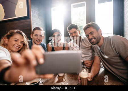 Diversi gruppi di amici prendendo selfie su smart phone. Giovani uomini e donne seduti al tavolo del bar e prendendo un autoritratto sul telefono cellulare. Foto Stock