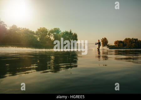 Colpo all'aperto dell'uomo Wakeboard sul lago al tramonto. Sci d'acqua sul lago dietro una barca. Foto Stock