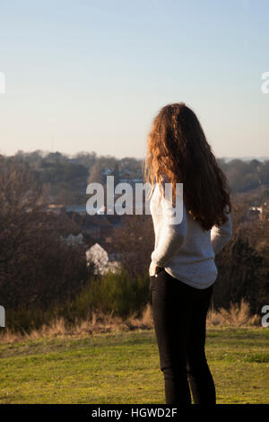 Giovane donna con capelli lunghi in piedi torna alla fotocamera, guardando in lontananza. Tre quarti di lunghezza. Foto Stock