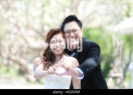 Ritratto di Allegro sposi abbracciando e mostra forma di cuore. Asian sposi in giardino Foto Stock