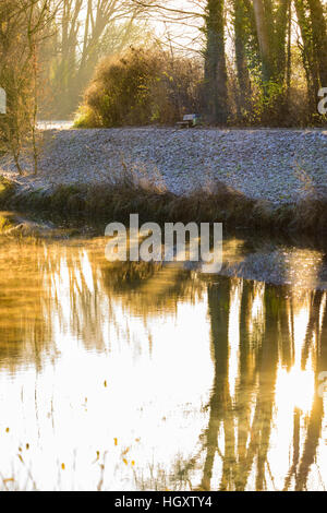 Golden misty autunno o inverno sunrise al fiume con acqua riflessi di alberi e una panchina nel parco a riposo a lato di una promenade Foto Stock