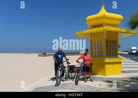 Due persone senior in marcia del ciclo con le biciclette da un giallo beach kiosk presso la spiaggia di Foz do Arelho sulla Costa d'argento del Portogallo Foto Stock