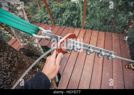 Mano trattiene l'anello di sicurezza di bloccaggio del cavo sull'imbracatura per camminare sulla parte superiore della struttura a ponte di legno. Foto Stock