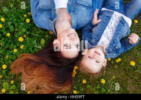 Madre e figlia sdraiati su un prato verde tra giallo di tarassaco. Ragazze vestite di denim giacche. La molla dell'umore. Aspetto familiare Foto Stock