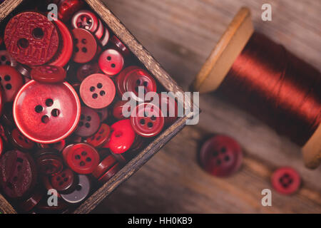 Una chiusura di una scatola di pulsanti rossi, con un rocchetto di filo rosso accanto a. Filtrato per avere un sbiadito finitura opaca e nostalgico Foto Stock