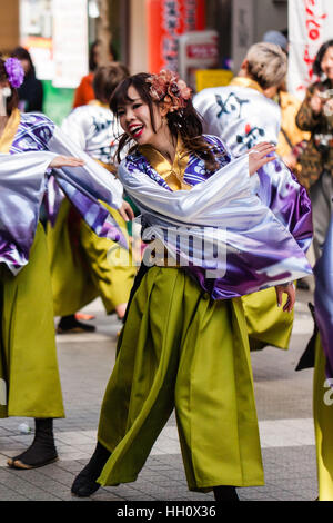 Yosakoi giapponese festival. Sorridente giovane donna, parte di un team, dancing in shopping arcade, ondeggiando le sue lunghe maniche malva. Fiore nei capelli. Foto Stock