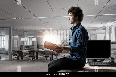 Giovane uomo in ufficio seduti sul tavolo con libro in mano. I supporti misti Foto Stock