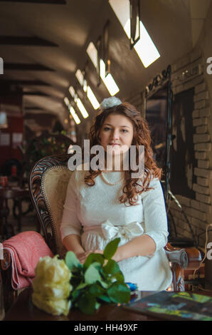 Il giovane è bella la ragazza in un abito bianco si siede su una sedia ad una grande finestra Foto Stock
