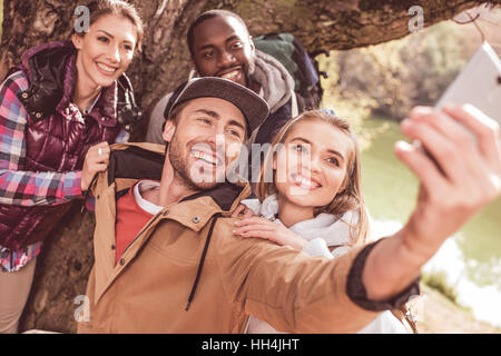 Un gruppo di giovani in piedi vicino al grande albero e tenendo selfie vicino tranquilla foresta fiume