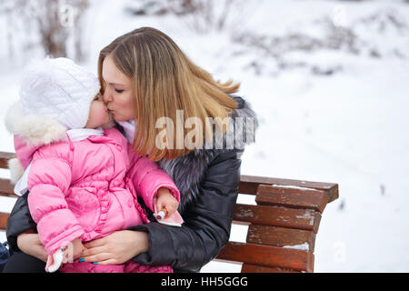 Madre figlia tiene tra le sue braccia e bacia il sul freddo all'esterno. Ella si è seduta sul banco di lavoro. Famiglia a piedi in un parco d'inverno. La felicità della famiglia. Foto Stock