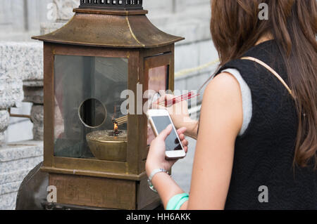 Donna con telefono cellulare luci bastoncini di incenso a Sik sik Yuen Wong Tai Sin Temple, Hong Kong Foto Stock