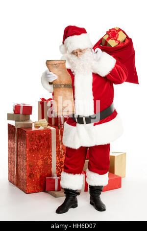 Felice Santa Claus in piedi con un mucchio di regali di Natale e un sacco di grandi dimensioni sul retro la lettura wishlist Foto Stock