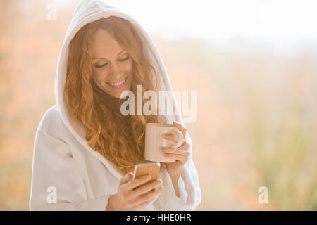 Donna sorridente in accappatoio con cappuccio texting con un telefono cellulare e di bere il caffè Foto Stock