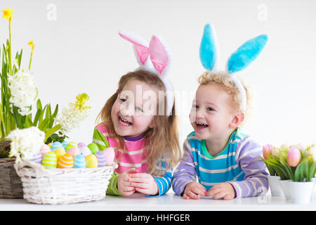 Dei bambini felici celebrare la Pasqua in casa. Un ragazzo e una ragazza indossando orecchie di coniglietto godendo di ricerca delle uova. I bambini a giocare con i colori delle uova Foto Stock