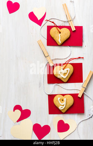 Cuori di carta e forma di cuore i cookie appeso su una stringa con mini vestiti pin su bianco tavola di legno Foto Stock