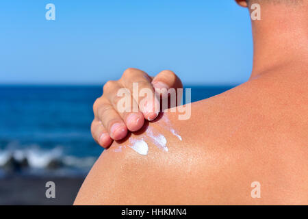 Primo piano di un giovane uomo caucasico visto da dietro sulla spiaggia l'applicazione di crema solare al suo indietro Foto Stock