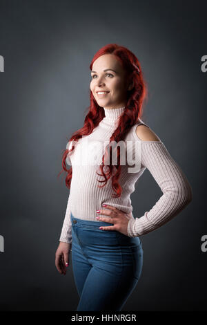 Tre quarti di lunghezza Ritratto di giovane donna con lunghi capelli rossi, sfondo grigio Foto Stock