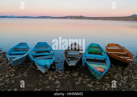 Cinque imbarcazioni a remi vuota sulla costa rocciosa del lago al tramonto Foto Stock