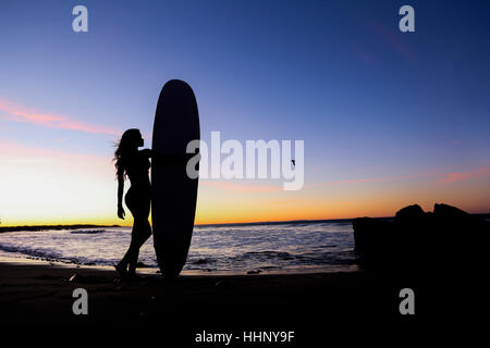 Silhouette di donna caucasica tenendo la tavola da surf in spiaggia Foto Stock