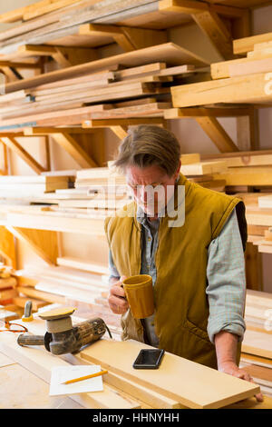 Falegname caucasica a bere caffè e utilizzando il cellulare in officina Foto Stock