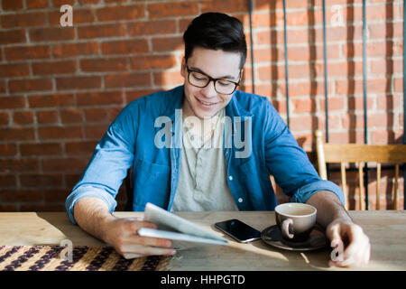 Giovane uomo sorridente, la lettura della carta, indossare occhiali e bere un caffè seduti ad un tavolo di legno in hipster cafe Foto Stock