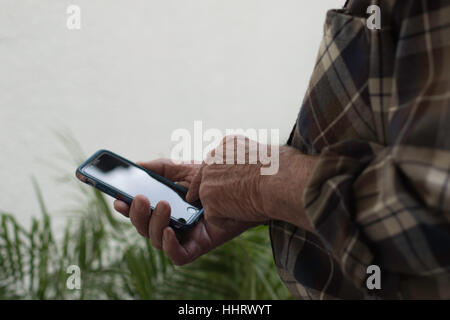 Mano d'uomo tenendo il cellulare in mano destra in piedi fuori sopra verdi piante con lo spazio negativo in alto a sinistra sullo sfondo puntando al pulsante home Foto Stock