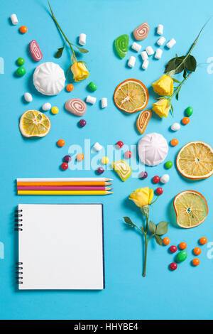 Marshmallows, candy, Jelly Beans, dolci , tazze secchi Arance e rose giallo che cade su un blocco note su uno sfondo blu. Spazi pubblicitari Foto Stock