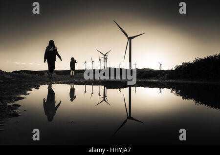Le turbine eoliche in una fattoria eolica con le sagome delle due ragazze si riflette sulla superficie dell'acqua al tramonto, Creta, Grecia Foto Stock