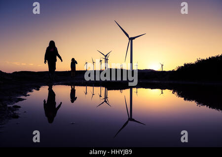 Le turbine eoliche in una fattoria eolica con le sagome delle due ragazze si riflette sulla superficie dell'acqua al tramonto, Creta, Grecia Foto Stock