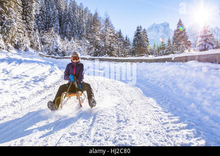 Allegro ragazza in sella a una slitta in discesa su una coperta di neve pista per slittino in un bianco inverno pieno di sole paesaggio di montagna Foto Stock