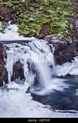 Una cascata circondata da ghiaccio e neve nelle Highlands di Islanda incorniciato terreni accidentati offre paesaggio panoramico epitomizing congelata di deserto. Foto Stock