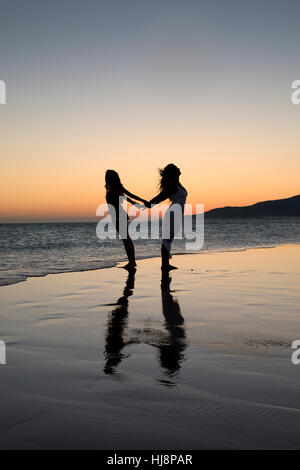 Silhouette di due donne tenendo le mani sulla spiaggia los Lances al tramonto, Tarifa, Cadice, Andalusia, Spagna Foto Stock