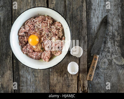 Preparazione di cotolette - materie carni macinate in un metallo smaltato ciotola, uovo e vintage coltello su un vecchio weathered tavolo in legno Foto Stock