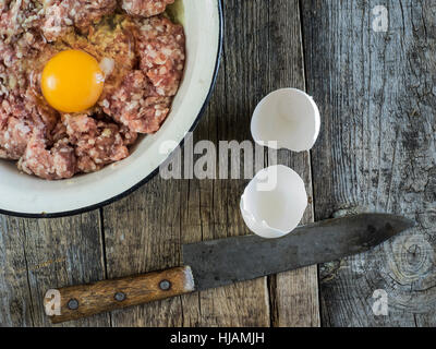 Preparazione di cotolette - materie carni macinate in un metallo smaltato ciotola, uovo e vintage coltello su un vecchio weathered tavolo in legno Foto Stock