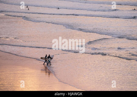 Surfers camminare nel mare come il sole tramonta su Fistral Beach in Newquay, Cornwall, Inghilterra. Foto Stock