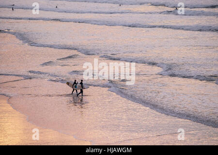 Surfers camminare nel mare come il sole tramonta su Fistral Beach in Newquay, Cornwall, Inghilterra. Foto Stock