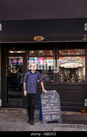 Ritratto di barman appoggiata contro l'happy hour board fuori casa pubblica finestra, Brooklyn, New York, Stati Uniti d'America Foto Stock