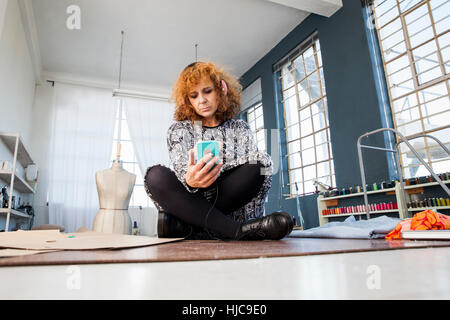 Femmina matura designer di moda con le gambe incrociate sul tavolo del workshop guardando smartphone Foto Stock