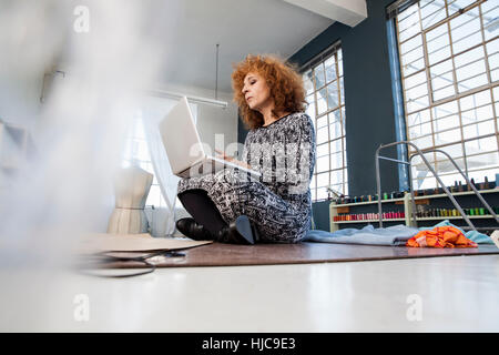 Femmina matura designer di moda con le gambe incrociate sul workshop tabella utilizzando il computer portatile Foto Stock